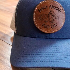 May include: A blue and white trucker hat with a brown leather patch that says "Shuck Around and Find Out" with a graphic of two oysters.