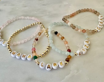 May include: Several beaded bracelets with white letter beads spelling out names like "Rosie", "Mama", "Lark", "Liza", and "Coral". The bracelets feature gold beads and various gemstone beads in shades of pink, brown, and green.