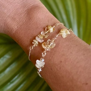 May include: Two delicate gold chain bracelets with small, irregular, light yellow and clear gemstone chips. The bracelets are layered on a person's wrist.