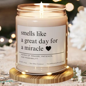 Puede incluir: Una vela de cera de soja encendida en un tarro de vidrio con el texto "smells like a great day for a miracle" y un símbolo de corazón. La vela está sobre un posavasos de madera. El texto "Notes of Prayer, Patience, and Possibility" está debajo.