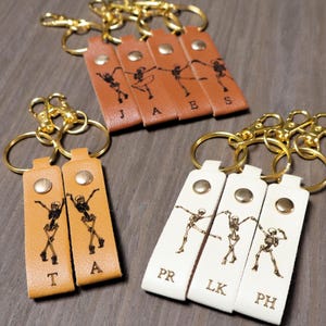 Può includere: Portachiavi in pelle marrone chiaro e bianco con finiture color oro. Ogni portachiavi presenta un disegno di ballerino scheletrico e una lettera. I portachiavi sono attaccati ad anelli color oro.