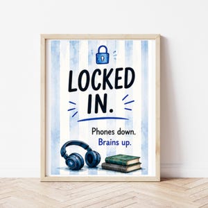 May include: A framed wall art print featuring a light blue and white vertical striped background. The text reads LOCKED IN. with a blue padlock icon above. Below, it says Phones down. Brains up. with illustrations of blue headphones and two books.