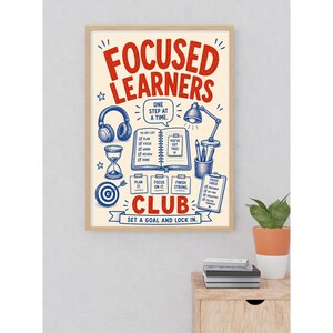 May include: A rectangular wall poster in a light wood frame featuring the red text FOCUSED LEARNERS CLUB and blue illustrations of headphones, an hourglass, a target, a desk lamp, a pencil holder, and an open notebook with a to-do list.