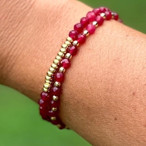 May include: Two beaded bracelets on a wrist. One bracelet features small, faceted, deep red beads. The other bracelet has small gold-colored beads. The bracelets are stacked together. The background is a soft green.