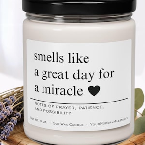 Puede incluir: Una vela de cera de soja blanca en un tarro de vidrio con tapa negra. La etiqueta dice "smells like a great day for a miracle" con un corazón negro. La vela es de 255 gramos y tiene un arreglo floral al lado.