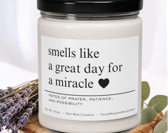 May include: A white soy wax candle in a glass jar with a black lid. The label reads "smells like a great day for a miracle" with a black heart. The candle is 9 oz. and has a floral arrangement next to it.