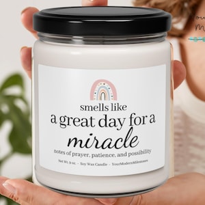 Puede incluir: Una vela de cera de soja blanca en un tarro de vidrio con tapa negra. La etiqueta presenta un arcoíris pastel y el texto "smells like a great day for a miracle". El texto adicional incluye "notes of prayer, patience, and possibility."