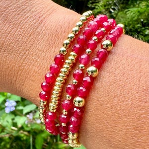 May include: A set of five bracelets featuring red gemstone beads and gold beads. The bracelets are stacked on a person's wrist.
