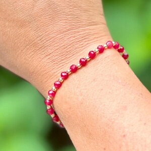 May include: A beaded bracelet featuring small, faceted, red gemstones and gold-colored beads. The bracelet is worn on a wrist, with a blurred green background.