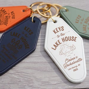 May include: Four keychains with the text "Keys to the Lake House" in brown lettering on a white, green, blue, and orange background. The white keychain has the text "Elaina's Bachelorette Lake George, NY 2025".