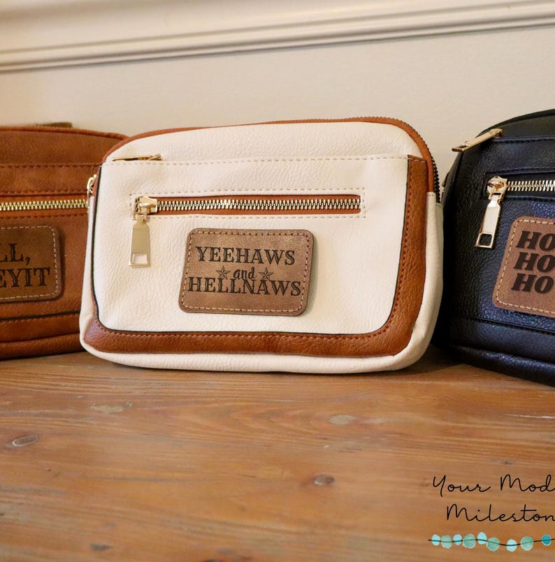 May include: Three small zippered bags with brown leather trim. The center bag is white with a brown leather patch that reads "Yeehaws and Hellnaws" with a star on each side of "and". The bag on the left is brown and the bag on the right is black.