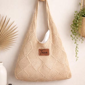 May include: A beige woven tote bag with a diamond pattern, hanging against a white wall. The bag has a wide opening and a leather patch with the name "Tara" on it. A white lining is visible inside the bag.