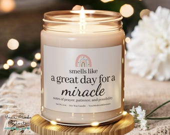 May include: A lit soy wax candle in a glass jar with a label that reads "smells like a great day for a miracle." The label features a rainbow graphic. The candle is on a wooden coaster.