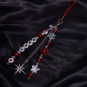 May include: A phone charm featuring a red cord, silver-tone metal chain, and beads. One strand includes white letter beads spelling CHAN, red round beads, and white heart beads. The other strand features a silver star charm and iridescent star beads.