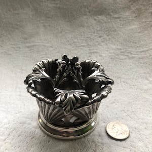 May include: Silver candle holder with a decorative leaf design. The holder is about the size of a quarter.