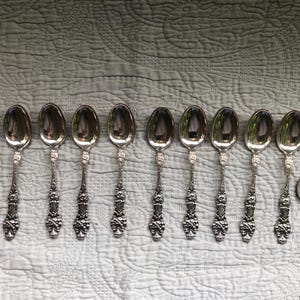 May include: Ten silver teaspoons with ornate handles. The spoons are arranged in a row on a white textured surface.