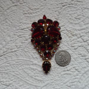 May include: A large, red rhinestone brooch with a teardrop-shaped pendant. The brooch is made of gold-toned metal and features a cluster of red rhinestones in a cascading design.