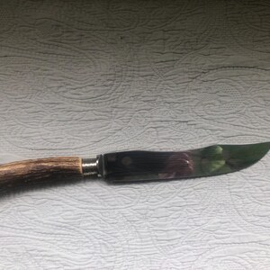 May include: A knife with a curved, polished blade and a handle made of brown antler. The blade has a dark, reflective surface with hints of green and purple. The handle is attached with a silver metal bolster.