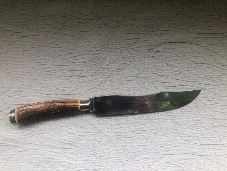 May include: A knife with a curved, polished blade and a handle made of brown antler. The blade has a dark, reflective surface with hints of green and purple. The handle is attached with a silver metal bolster.