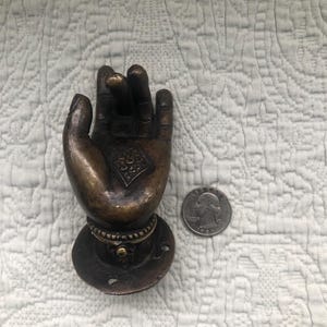 May include: A bronze hand door knocker with intricate details. The hand is in a gesture of giving or receiving. The knocker is attached to a round base with three holes.