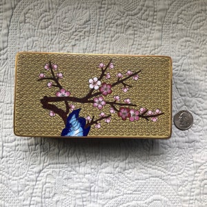 May include: A rectangular gold-colored box with a floral design. The box features a stylized cherry blossom branch with pink flowers and a blue and white bird on a gold background with a repeating pattern.