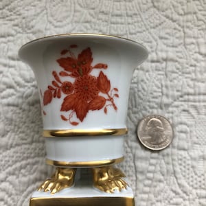 May include: A white porcelain vase with a floral design in shades of orange and gold. The vase has a square base and is decorated with gold trim. The vase is approximately 5 inches tall.