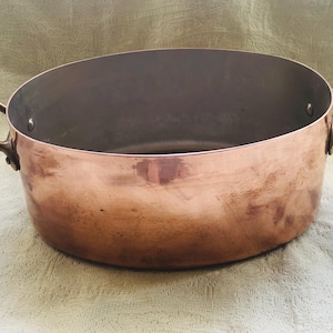 May include: A copper cooking pot with two brass handles. The pot has a wide, oval shape and a shiny, reddish-brown exterior. The interior is a darker, metallic color. The handles are curved and attached to the sides of the pot.