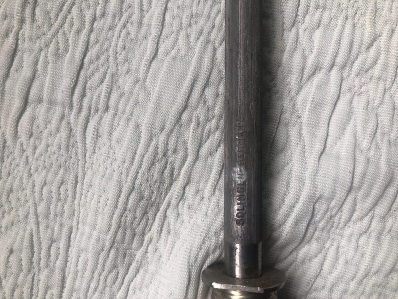 May include: Close-up of a metal knife sharpener with the text "SOLINGEN GERMANY" etched on the side. The sharpener has a dark, textured handle and a metal base. The background is a white, textured fabric.