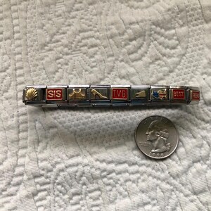 May include: A silver charm bracelet with gold and red accents, featuring various charms with words and symbols. A quarter is next to the bracelet for size comparison. The charms include the words "SIS", "TVB", and "BEST".