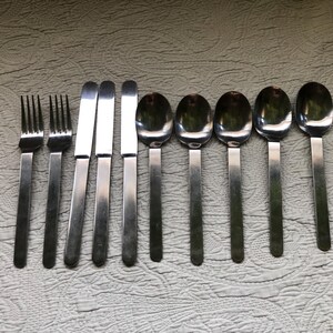 May include: A set of stainless steel flatware, including forks, knives, and spoons, arranged on a textured surface. The silverware has a simple, modern design with long, rectangular handles and rounded spoon bowls. The set includes three forks, three knives, and four spoons.