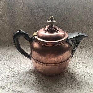 May include: A copper teapot with a dark handle and spout. The lid is topped with a gold-colored finial. The spout and handle have decorative metal accents. The teapot is sitting on a white patterned surface.