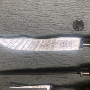May include: A knife with a decorative blade featuring an etched design and a brown handle. The blade has a silver finish. A quarter is next to the knife for scale. The knife is part of a set.