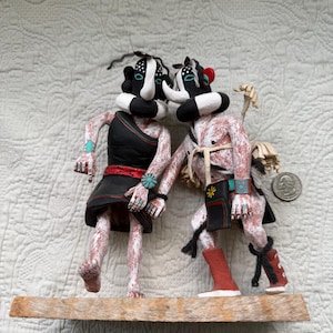 May include: Two handcrafted Native American Kachina dolls. One doll wears a black dress and the other wears a red and black outfit. Both dolls have black and white masks and are standing on a wooden shelf. The dolls are on a white quilted background.