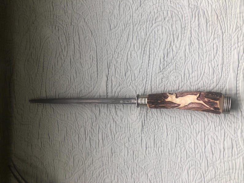 May include: A steel knife sharpener with a decorative handle. The handle is made of brown wood and features a carved image of a deer. The handle is approximately 5 inches long.