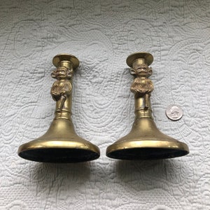 May include: Two antique brass candlesticks with a whimsical monkey design. Each candlestick features a wide base, a slender stem, and a small platform for holding a candle. The monkey figure is intricately detailed. A quarter is next to the candlesticks.