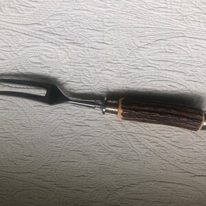 May include: A stainless steel carving fork with a two-pronged design. The handle is made of brown, textured material, possibly wood or horn, with silver-colored metal accents. The fork is designed for serving meat.