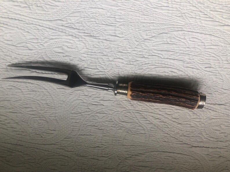 May include: A stainless steel carving fork with a two-pronged design. The handle is made of brown, textured material, possibly wood or horn, with silver-colored metal accents. The fork is designed for serving meat.
