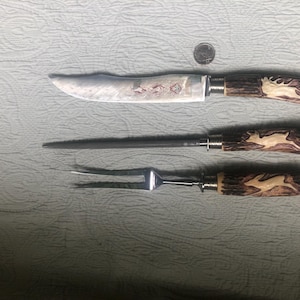 May include: A carving set with a knife, a sharpening steel, and a fork. The handles are brown with a deer carving. The knife blade has an etched design. A quarter is next to the set for scale.