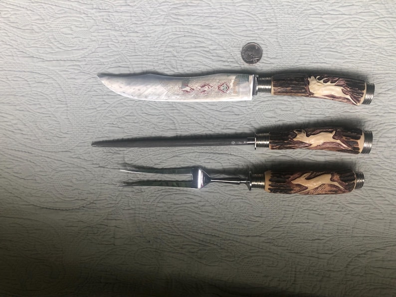May include: A carving set with a knife, a sharpening steel, and a fork. The handles are brown with a deer carving. The knife blade has an etched design. A quarter is next to the set for scale.
