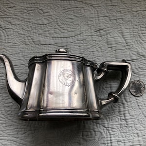 May include: Silver teapot with a decorative design and a crest on the front. The crest features a flag with a star.