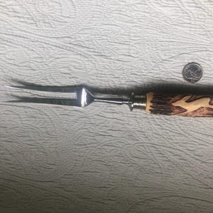 May include: A carving fork with a stainless steel head and a handle made of carved wood. The handle features a light-colored, carved image of a leaping deer. The fork is approximately 12 inches long.