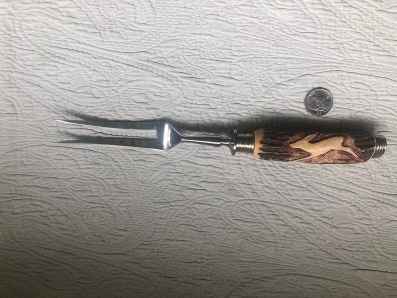 May include: A carving fork with a stainless steel head and a handle made of carved wood. The handle features a light-colored, carved image of a leaping deer. The fork is approximately 12 inches long.