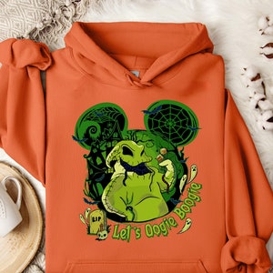 May include: Orange hoodie featuring a green Oogie Boogie design with a haunted house, bats, and the text "Let's Oogie Boogie." The design includes a tombstone and dice. The hoodie is made of a soft material.