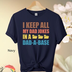May include: Navy blue t-shirt with the text "I KEEP ALL MY DAD JOKES IN A DAD-A-BASE" in brown, orange, yellow, and blue. The shirt has a knot at the bottom and a Trendy Ink Fashion logo.