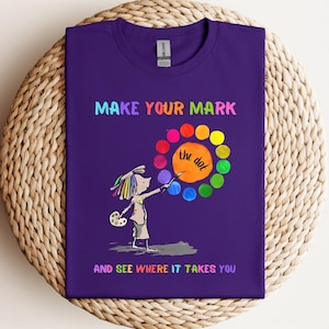 May include: Purple t-shirt with a colorful paint palette and a cartoon character holding a paintbrush. The text reads "Make your mark the dot and see where it takes you."
