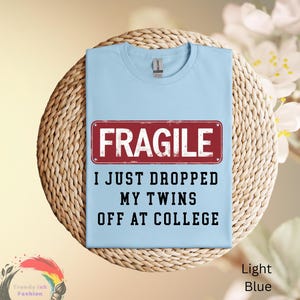 May include: Light blue t-shirt with the text "FRAGILE I JUST DROPPED MY TWINS OFF AT COLLEGE" in black and a red rectangle. The shirt is displayed on a woven, circular mat.