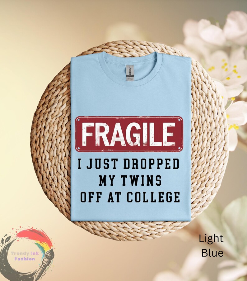 May include: Light blue t-shirt with the text "FRAGILE I JUST DROPPED MY TWINS OFF AT COLLEGE" in black and a red rectangle. The shirt is displayed on a woven, circular mat.
