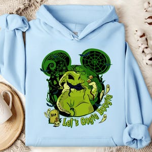 May include: Light blue hoodie featuring a green Oogie Boogie graphic with a haunted house, bats, and spiderwebs. The text "Let's Oogie Boogie" is also displayed. The design is set against a Mickey Mouse ear silhouette.