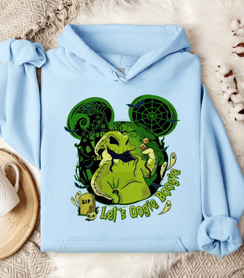 May include: Light blue hoodie featuring a green Oogie Boogie graphic with a haunted house, bats, and spiderwebs. The text "Let's Oogie Boogie" is also displayed. The design is set against a Mickey Mouse ear silhouette.