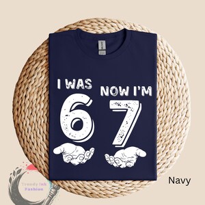 May include: Navy blue t-shirt with the text "I WAS 67 NOW I'M" in white, along with the number 67 and two open hands. The shirt is displayed on a woven mat.